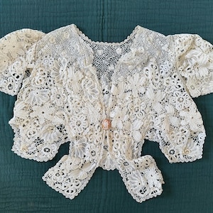 May include: Ivory-colored lace bolero jacket with floral and geometric patterns. The jacket features short sleeves and a decorative clasp. The intricate lace design is visible throughout the garment, creating a delicate and vintage aesthetic.