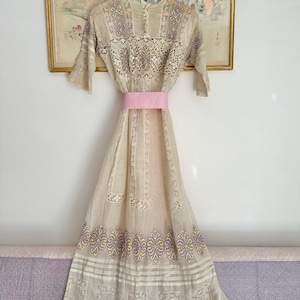 Può includere: Un abito vintage beige con pizzo e ricami floreali intricati. L'abito presenta una cintura rosa ed è appeso a una gruccia di legno. L'abito ha una gonna lunga con motivi decorativi ed è esposto contro un muro con un'opera d'arte incorniciata.