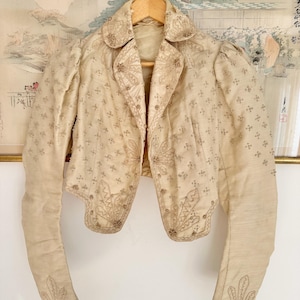 Könnte beinhalten: Eine cremefarbene, kurz geschnittene Jacke mit langen Ärmeln, Puffschultern, einem Kragen und aufwendigen Stickereien im gleichen Farbton. Das Kleidungsstück ist mit kleinen, sich wiederholenden Blumenmustern und Ziernähten an den Rändern versehen.