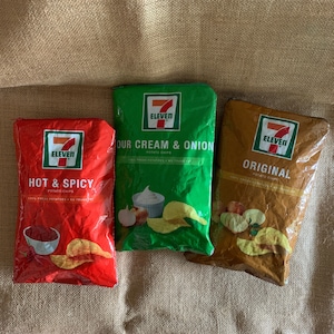 May include: Three bags of 7-Eleven potato chips. The bags are red, green, and brown. The red bag says "Hot & Spicy", the green bag says "Our Cream & Onion", and the brown bag says "Original".