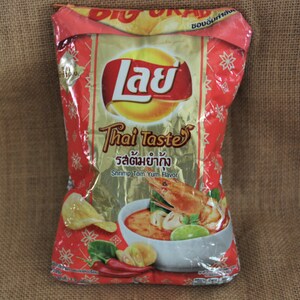 May include: A red and gold bag of Lay's Thai Taste potato chips with a picture of a bowl of shrimp tom yum soup. The bag has the text "Lay's Thai Taste" and "Shrimp Tom Yum Flavor" in English and Thai.