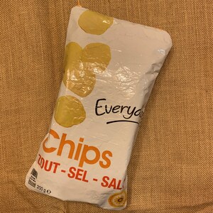 Può includere: Un sacchetto bianco di patatine con la scritta "Chips" in arancione e "Everyday" in nero. La confezione presenta illustrazioni di patatine gialle e la scritta "ZOUT - SEL - SAL". La confezione contiene 200g di patatine.