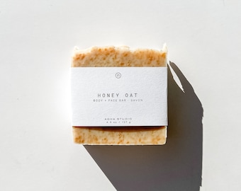 May include: A white bar of soap with a label that reads "Honey Oat Body + Face Bar Savon Agha Studio 4.8 oz / 137 g".
