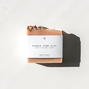 May include: A rectangular bar of French pink clay soap with a white label. The label reads "FRENCH PINK CLAY BODY + FACE BAR SAVON" and "AGHA STUDIO 4.5 oz / 127 g". The soap is a light pink color and has dried flower petals on top.