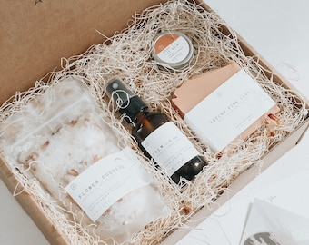 May include: A brown cardboard box filled with shredded paper and a gift set of bath and body products. The products include a small jar of lip balm labelled "Body Lip Balm", a bottle of spray labelled "Calm", a bag of bath salts labelled "Flower Goddess", and a bar of soap labelled "French Pink Clay".