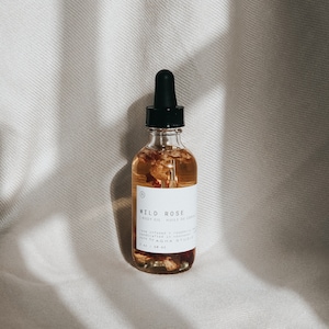 Peut inclure: Une bouteille en verre transparent avec un compte-gouttes noir rempli d'un liquide ambré clair et de pétales de rose. L'étiquette de la bouteille indique "WILD ROSE BODY OIL HUILE DE CORPS rose infused + raspberry seed oil + vitamin e made by AGHA STUDIO 2 oz / 60 ml"