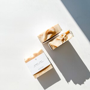 May include: Three bars of soap, two are stacked on top of each other, and one is laying flat. The soap is a light brown color and has a white label that says "Honey Oat Body + Face Bar Soap" and "AURA STUDIO 4 oz / 113 g".