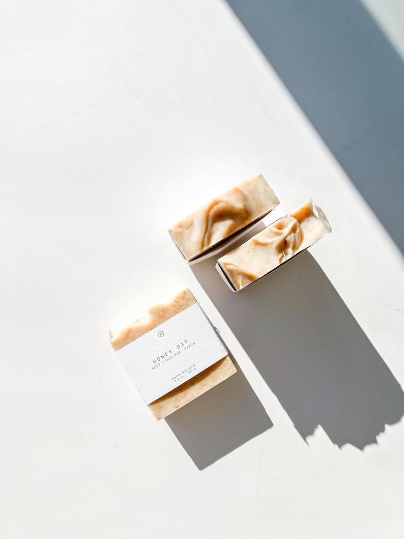May include: Three bars of soap, two are stacked on top of each other, and one is laying flat. The soap is a light brown color and has a white label that says "Honey Oat Body + Face Bar Soap" and "AURA STUDIO 4 oz / 113 g".
