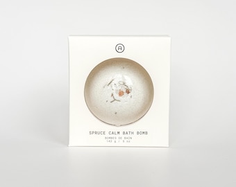 Puede incluir: Una caja blanca con un recorte circular que muestra una bomba de baño. La bomba de baño está moteada con acentos marrones claros y verdes. El texto en la caja dice "Spruce Calm Bath Bomb" y "142 g / 5 oz".