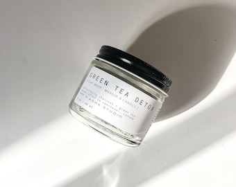 May include: A small glass jar with a black lid containing a green tea detox clay mask. The label reads "GREEN TEA DETOX | CLAY MASK - MASQUE À L'ARGILE | Detoxifying charcoal + green tea handcrafted in Vancouver, Canada by AGHA STUDIO | 1 oz / 68 ml"