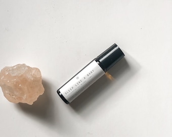 Puede incluir: Un pequeño frasco de vidrio transparente con un aplicador de bola negro. El frasco está etiquetado con "SLEEP LIKE A BABY | ESSENTIAL OIL BLEND (5ML)".