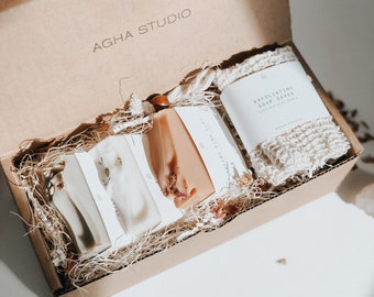 May include: A brown cardboard box with a white label that says "AGHA STUDIO" is open and filled with natural bath products. The box contains two bars of soap, a small bag of dried flowers, and a white exfoliating soap saver with the text "EXFOLIATING SOAP SAVER 100% NATURAL BAMBOO AGHA STUDIO".