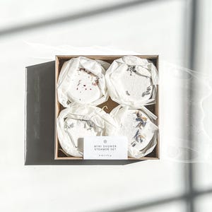 May include: A set of four mini shower steamers in a white box with a label that reads "Mini Shower Steamer Set 4 oz / 113 g". The steamers are wrapped in white tissue paper and have dried flowers and herbs inside.