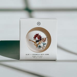 May include: A white square cardboard box with a circular cutout displaying a round white bath bomb decorated with dried flower petals. The text on the box reads ALOE + COCOMILK BATH BOMB, BOMBES DE BAIN, 142 g / 5 oz, and features a small circular logo.