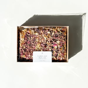 May include: A clear plastic box filled with dried flower petals for a bath tea. The label reads "Flower Kind Bath Tea Tisané de Bain 1.8 oz / 51 g".