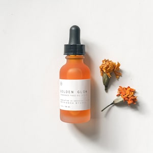 May include: A clear glass bottle with a black dropper top filled with a golden orange liquid. The bottle is labelled "GOLDEN GLOW RADIANCE FACE OIL SERUM" and "AGHA STUDIO". The bottle is sitting on a white surface with two dried orange flowers.