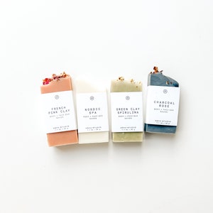 May include: Four bars of soap with different scents and ingredients. The bars are arranged from left to right: French Pink Clay, Nordic Spa, Green Clay Spirulina, and Charcoal Rose. Each bar has a label with the name of the scent and ingredient, as well as the brand name "AGHA STUDIO".