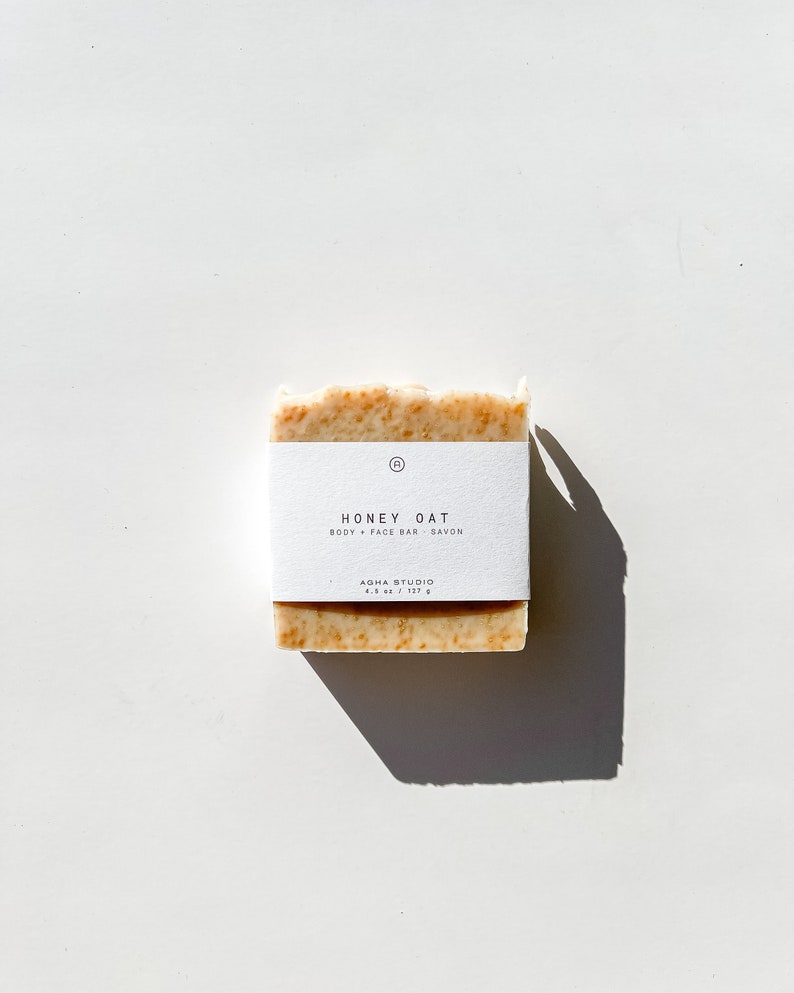May include: A white bar of soap with a label that reads "Honey Oat Body + Face Bar Savon Agha Studio 4.8 oz / 137 g".