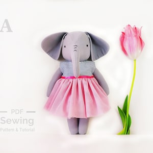 May include: A grey fabric elephant doll wearing a pink tulle skirt and a blue and white striped shirt. The doll is standing on a white surface next to a pink tulip.