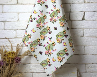 Op de afbeelding: Een witte keukendoek met een bloemenpatroon met roze bloemen, groene bladeren en gele vogels in kooien. De handdoek hangt aan een haak.