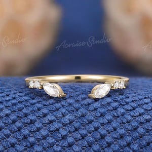 May include: A gold-colored ring with a unique open design. The ring features two marquise-cut white gemstones at the open ends and small round white gemstones along the band. The ring is set against a textured blue background.
