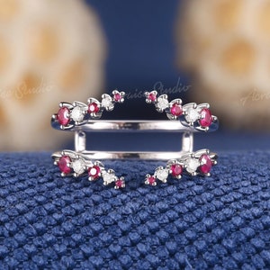 Puede incluir: Un delicado anillo de plata con un diseño dividido, que presenta una fila de pequeñas piedras preciosas rojas y blancas a cada lado. El anillo está diseñado para ser usado como una banda de boda o un anillo de compromiso.