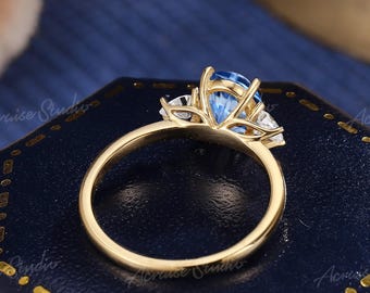 Puede incluir: Un anillo de oro con una piedra preciosa azul redonda central flanqueada por dos piedras claras más pequeñas. El engaste está elevado con un delicado diseño de cesta de metal. El anillo está colocado sobre una caja de presentación hexagonal de color azul oscuro con borde dorado.