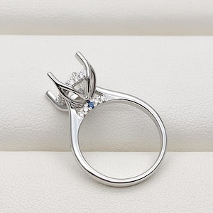 May include: A silver-toned metal ring setting featuring a high prong mount designed to hold a central gemstone. The band includes small clear accent stones and a single small blue stone embedded on each side of the setting near the prongs.