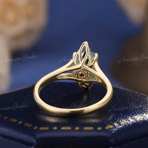 May include: A gold ring featuring a marquise-cut green gemstone held in a prong setting. The band has a split shank design that connects to the stone setting, which includes a small, circular red accent stone embedded at the base of the setting.
