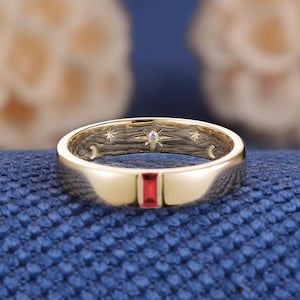 May include: A gold band ring featuring a single rectangular red gemstone set into the outer surface. The interior of the band displays a textured finish with engraved celestial motifs, including stars and a crescent moon, and a small embedded diamond.