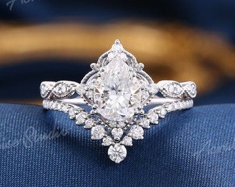 Puede incluir: Un anillo de compromiso de diamante de forma pera con una banda delicada y adornada. El anillo presenta una piedra central de forma pera rodeada de diamantes más pequeños, creando un look de inspiración vintage.