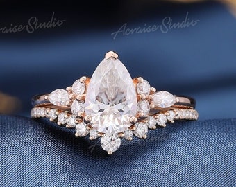 Puede incluir: Un anillo de compromiso de diamante de forma pera engastado en oro rosa con un halo de diamantes más pequeños. El anillo está sobre un fondo de tela azul.