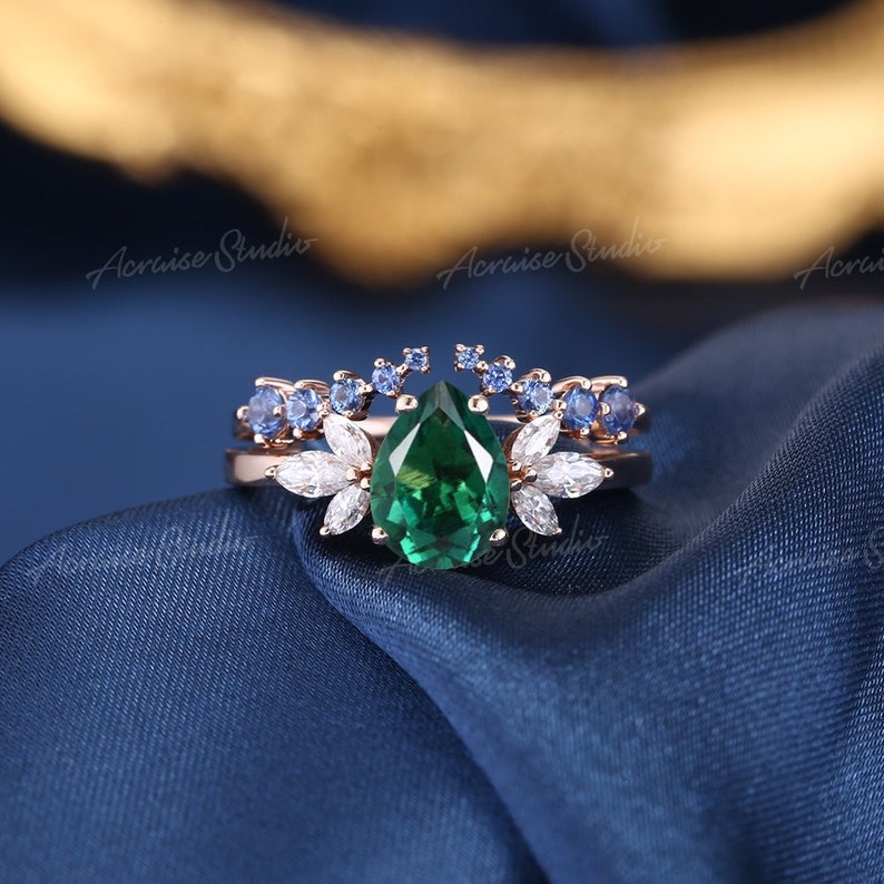May include: A pear-shaped emerald green gemstone ring with a delicate floral design featuring smaller white and blue gemstones. The ring is set in rose gold.