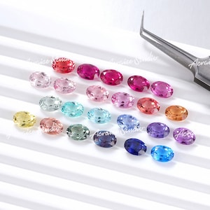 Puede incluir: Surtido de piedras preciosas ovaladas en varios colores, incluyendo amarillo, rosa, azul, verde, rojo y morado. Las piedras preciosas están dispuestas en filas sobre una superficie blanca.