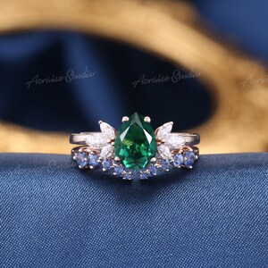 May include: A pear-shaped emerald green gemstone ring with a halo of smaller white gemstones. The ring is set on a rose gold band and is paired with a matching band featuring blue sapphires.