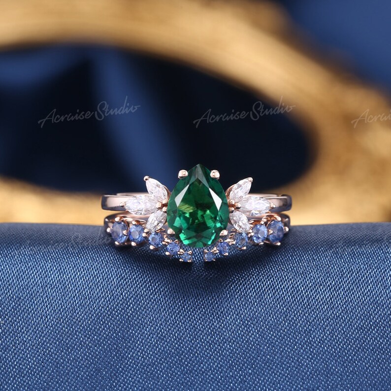 May include: A pear-shaped emerald green gemstone ring with a halo of smaller white gemstones. The ring is set on a rose gold band and is paired with a matching band featuring blue sapphires.
