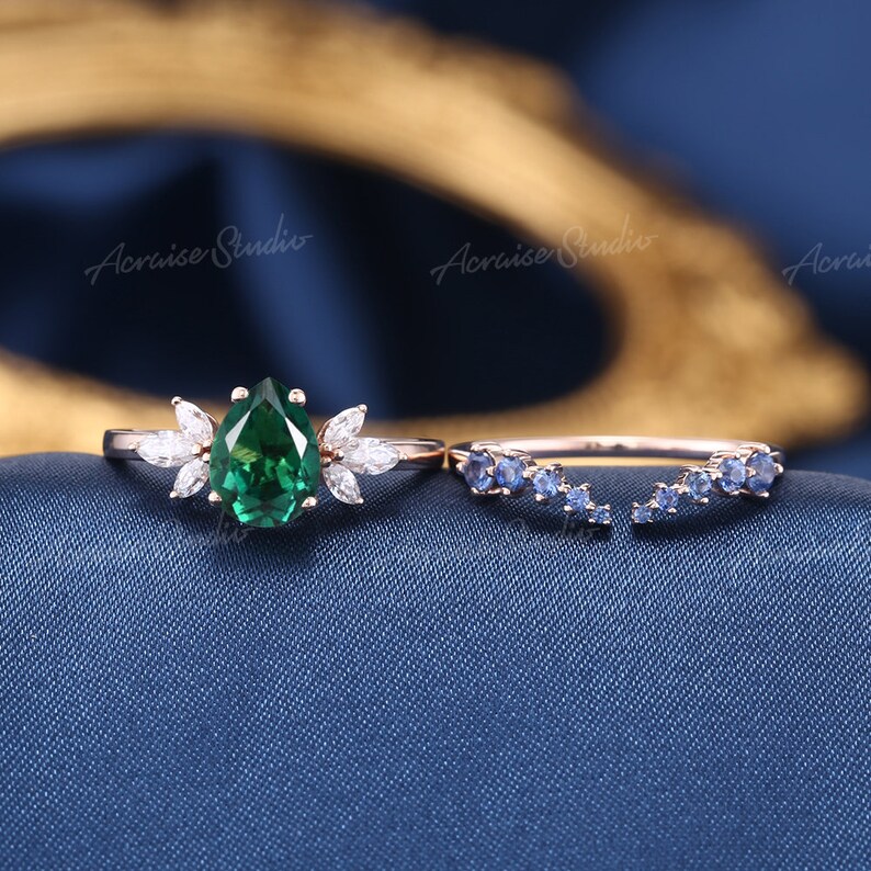 May include: A pear-shaped emerald engagement ring with a delicate floral design and a matching sapphire band. The ring is set in rose gold and features a cluster of small diamonds.