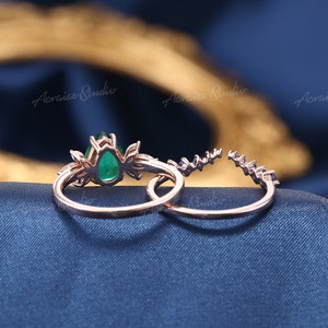 May include: A close-up of a rose gold engagement ring set with a pear-shaped emerald gemstone. The ring is accented with smaller diamonds and is set on a delicate band.