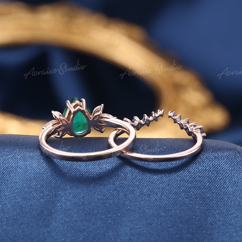May include: A close-up of a rose gold engagement ring set with a pear-shaped emerald gemstone. The ring is accented with smaller diamonds and is set on a delicate band.