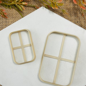 May include: Two beige rectangular cookie cutters with a windowpane design. The cutters are made of plastic and are set on a white surface. The cutters are different sizes, one is smaller than the other. The background is a neutral color.