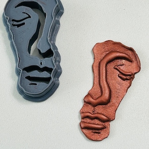 May include: Two face-shaped objects. One is a gray plastic cookie cutter with a layered design. The other is a reddish-brown, textured, face-shaped object. Both have closed eyes, a nose, and a mouth.