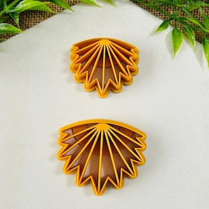 May include: Two yellow plastic cookie cutters in a fan or sunburst design. The cutters have a scalloped edge and radiating lines. The cutters are on a white surface with green foliage in the background.