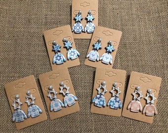 May include: Seven pairs of dangle earrings displayed on brown cardboard cards. Each pair features a miniature sweater charm paired with a Star of David or snowflake charm in various shades of blue, white, and light orange with patterned designs.