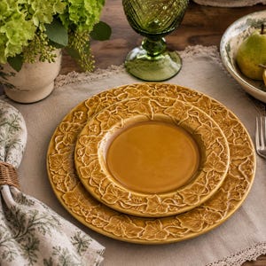 May include: Two mustard-yellow ceramic plates with a leaf design are set on a beige tablecloth. A silver fork, a green glass, and a bowl of pears are also on the table. A napkin with a woven ring and a vase of green flowers complete the dining setting.