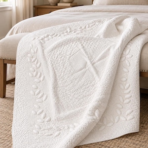 May include: A solid white quilt featuring intricate stitching patterns, including a central diamond motif with radiating lines, a textured stippled background, and a decorative border composed of repeating leaf-like shapes along the edges.