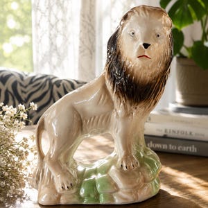 May include: A ceramic lion figurine standing on a green rock base. The lion has a cream-colored body, a dark brown mane, and painted facial features including black eyes, a black nose, and a thin red line for the mouth, with a glossy finish.