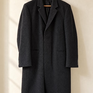 May include: A charcoal gray overcoat featuring a notched lapel, a single-breasted front with a hidden button placket, long sleeves with three small buttons at each cuff, and two side welt pockets, hanging on a wooden hanger.