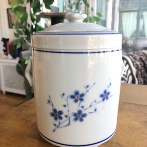 May include: White ceramic jar with a lid, adorned with blue floral patterns and a blue stripe. The jar has two small handles and a decorative knob on the lid. The jar is sitting on a wooden surface.