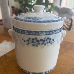 May include: A white ceramic lidded jar featuring a blue and light blue floral pattern around the rim and body. The jar has two small side handles and a matching lid with a central knob. The design includes delicate blue flowers and leaves.