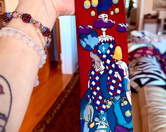 May include: A red necktie featuring a whimsical jester design. The tie showcases a jester figure with a blue and white diamond pattern, holding a globe. The design includes clouds, yellow orbs, and other stylized figures. The tie is held up by a person.